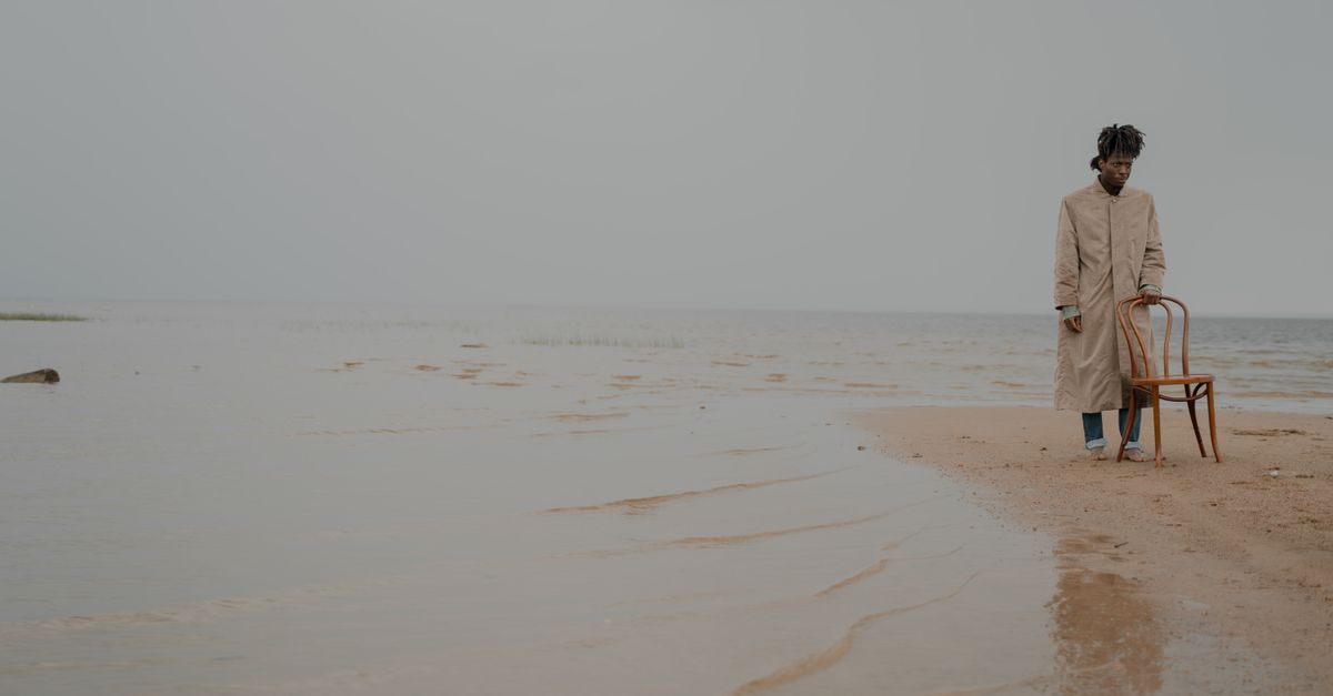 A man in a coat stands contemplatively on a serene beach beside a wooden chair.