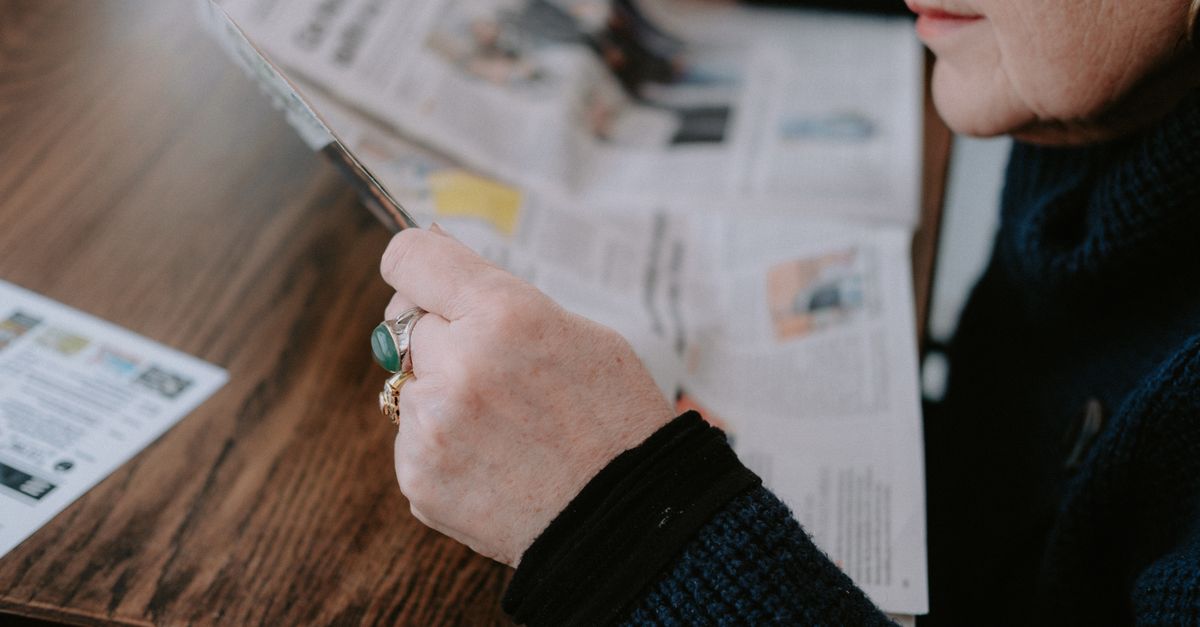 A woman reads a newspaper at a wooden table, focusing on leisure and information.