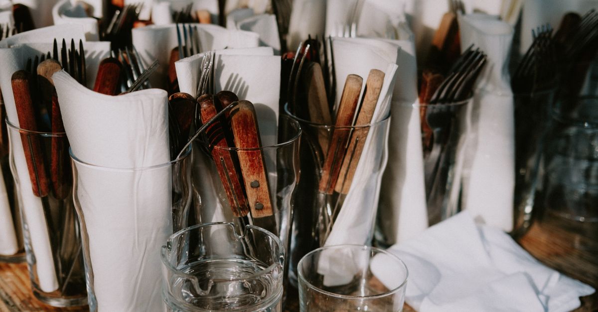 A collection of rustic wooden and metal cutlery in glass cups, neatly arranged with napkins on a wooden table.