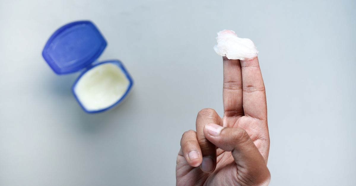 A hand holding petroleum jelly on fingers, next to an open container. Skin care concept.