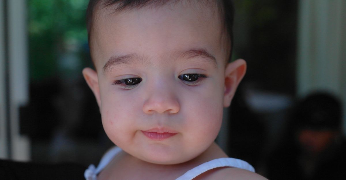 Close-up of a serene baby with a thoughtful expression, wearing a white dress indoors.
