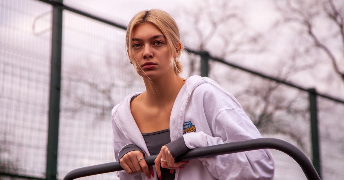 Young blonde woman posing in a white jacket at an outdoor gym setting.