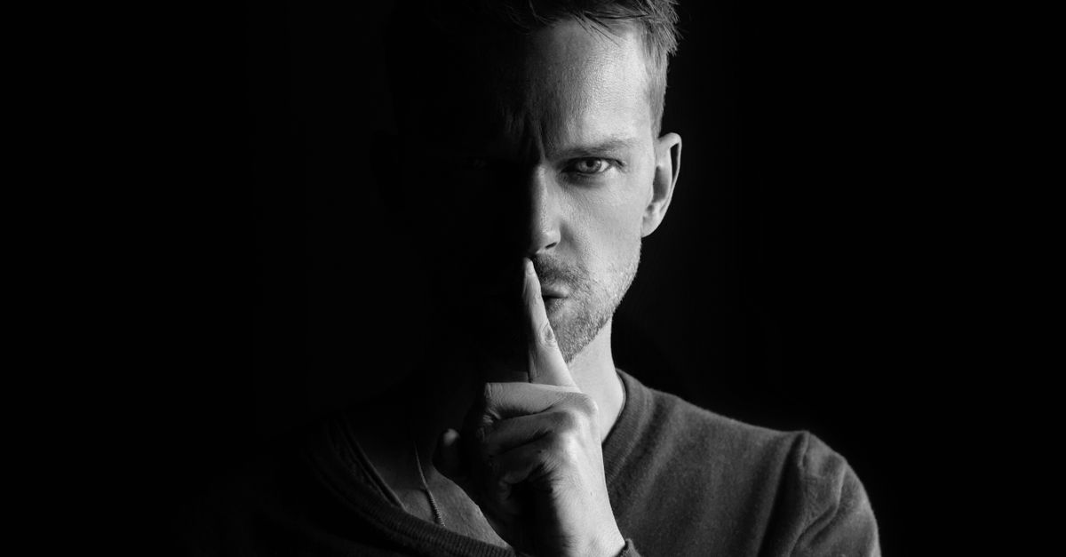 Black and white portrait of a man gesturing for silence, highlighting mystery.