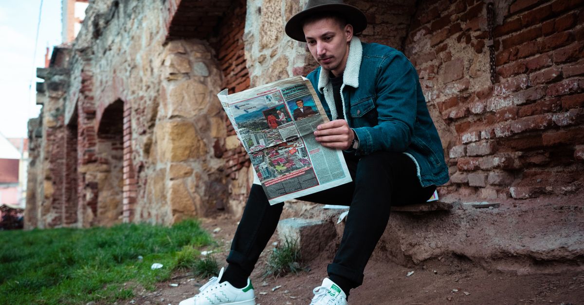 Young man in casual wear reading a newspaper against an ancient brick wall outdoors.
