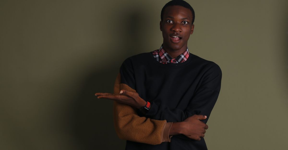 A young man in casual fashion poses expressively against a plain background.