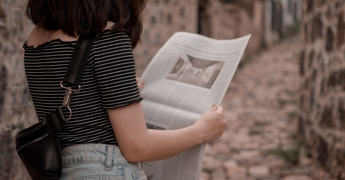 A woman stands outdoors on a cobblestone path reading a newspaper, embodying leisure and style.