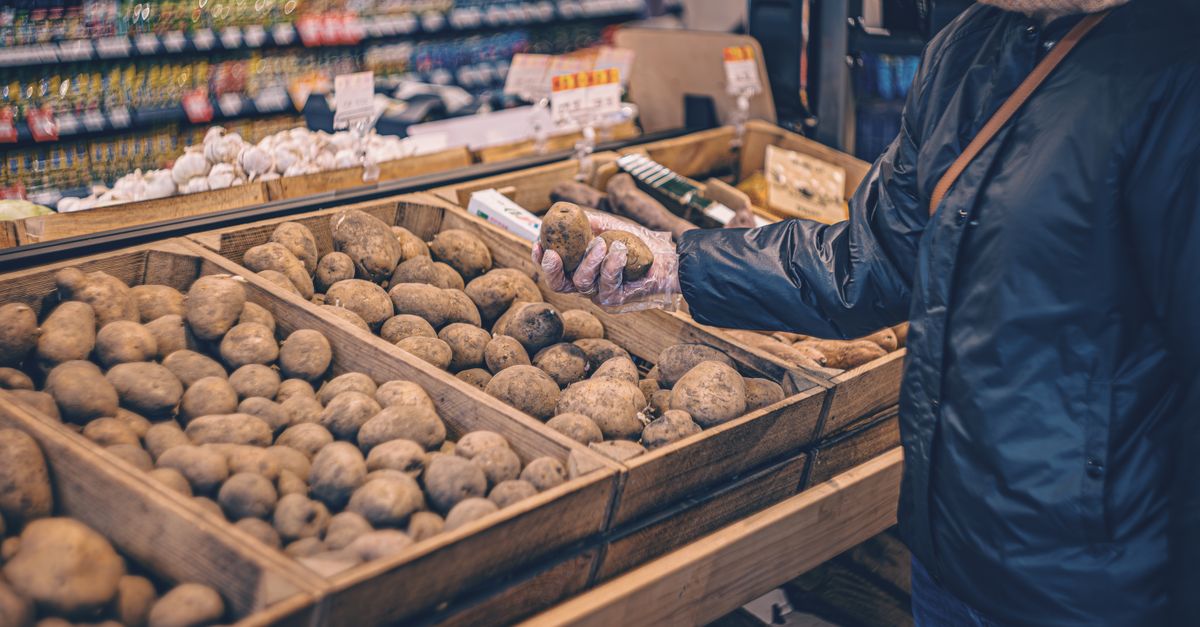 A shopper picks potatoes from a wooden crate in a grocery store produce section.