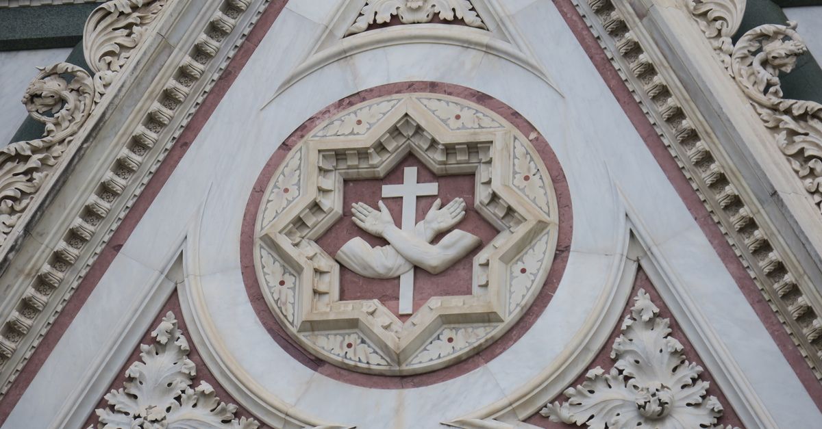 Close-up of a decorative architectural sculpture featuring a cross and hands, highlighting intricate carvings.