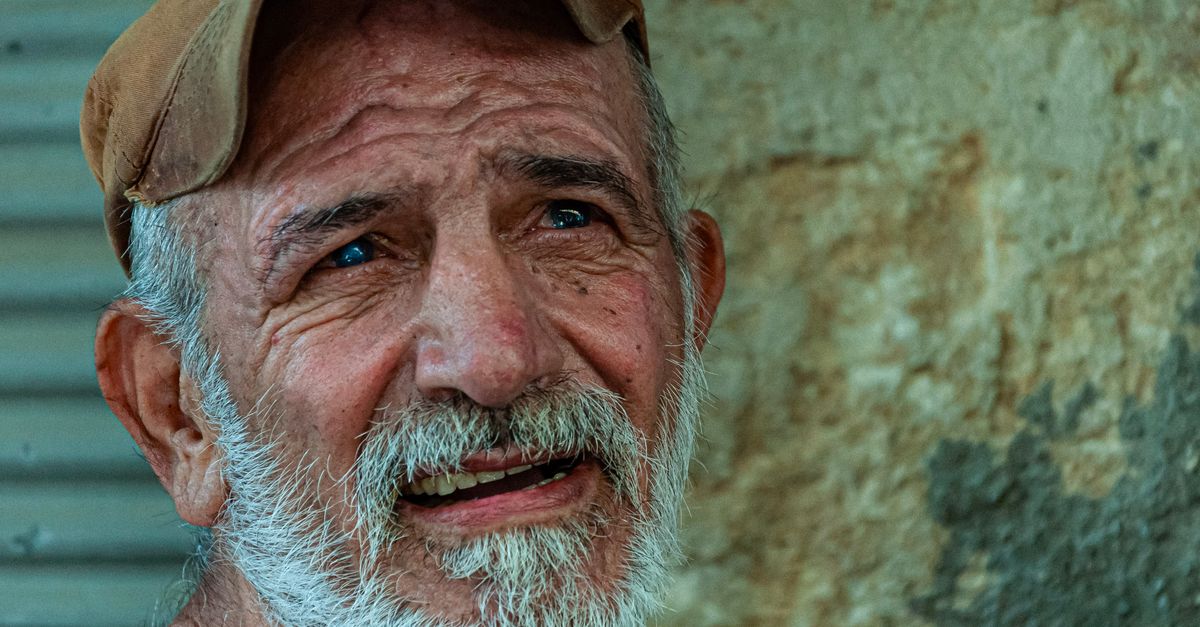 Portrait of an elderly man wearing a cap, smiling thoughtfully against a textured outdoor wall.