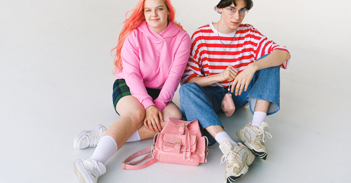 From above of full body informal teenager couple wearing colorful clothes sitting on floor with camera and backpack against white background and looking at camera