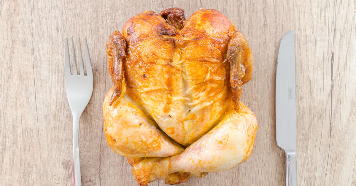 Top view of a perfectly roasted chicken placed between fork and knife on a wooden table.
