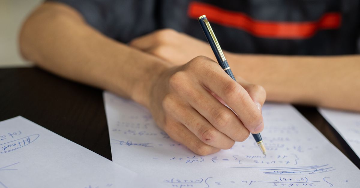 Unrecognizable smart male student with pen  taking notes while solving equations at table during lesson in classroom on blurred background
