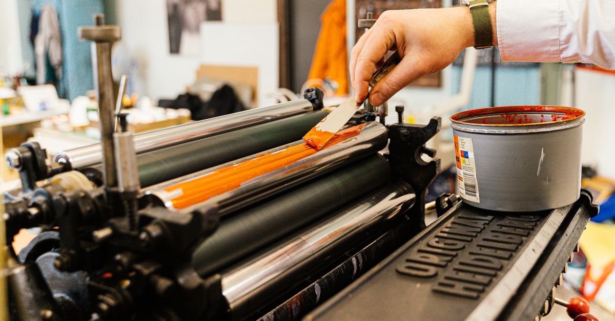 A detailed view of a hand applying orange ink to a vintage printing press roller indoors.