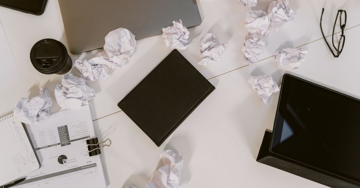 Top view of a cluttered office desk with crumpled papers, laptop, and eyeglasses.