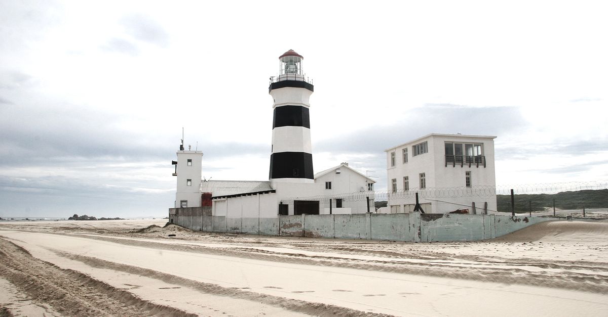 Exterior of striped beacon tower and houses located behind fence on sandy seacoast on cloudy gray day
