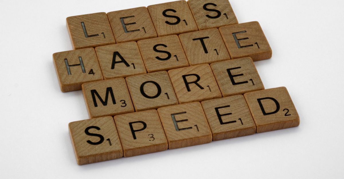 Wooden scrabble tiles forming the phrase 'Less Haste More Speed' on a white background.