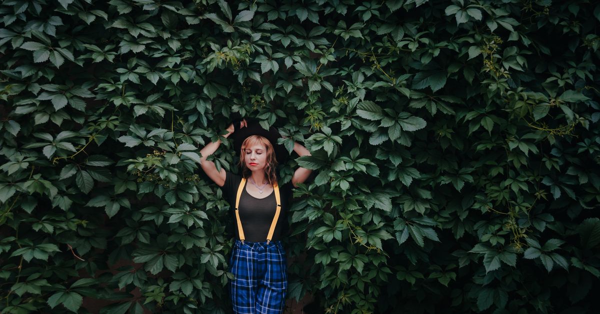 A fashionable woman with raised arms poses in front of dense greenery, exuding a stylish vibe.