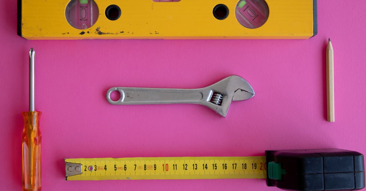 Flat lay of carpentry tools including level, wrench, screwdriver, measuring tape on pink background.