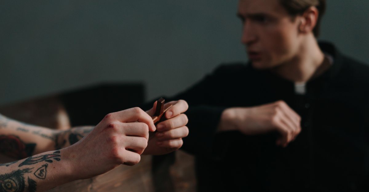 A tattooed person holds a crucifix while facing a priest, suggesting themes of faith and repentance.