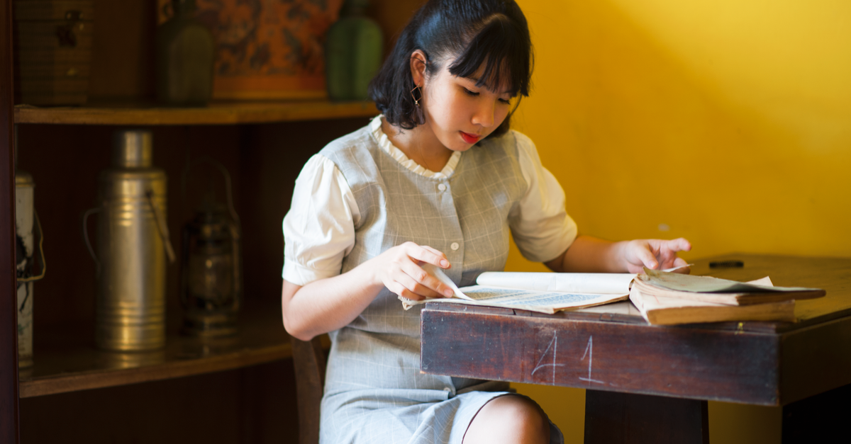 Asian woman engrossed in reading at desk with bookshelf and vintage decor in background.