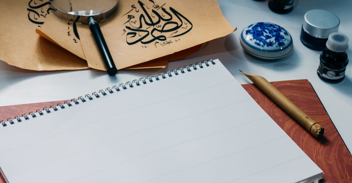 Creative calligraphy setup with Arabic script papers, magnifying glass, ink bottles, and blank notepad.
